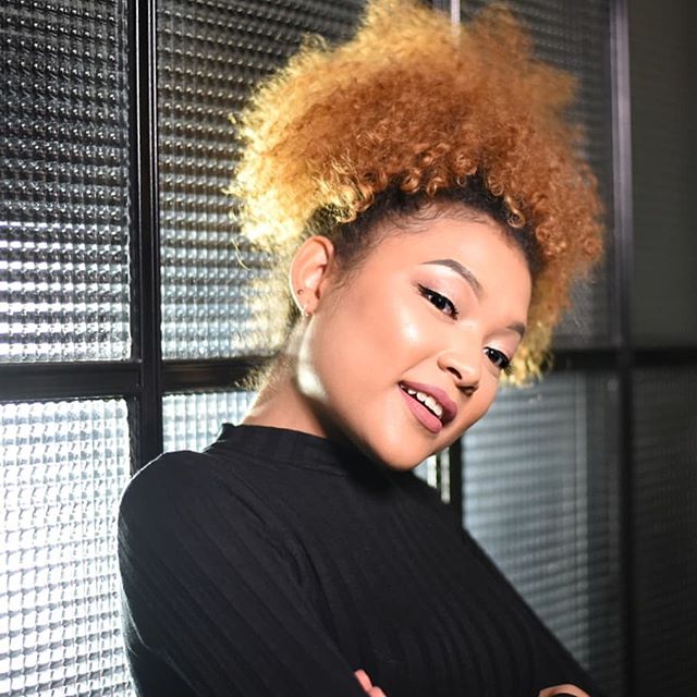 A young woman smiles confidently with her arms crossed, showcasing a vibrant hairstyle.