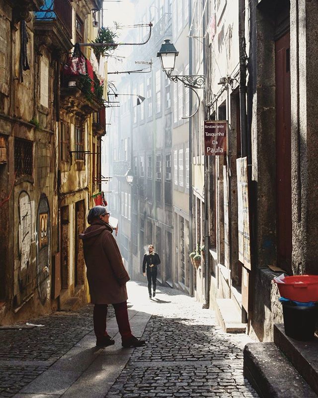 A woman stands on a cobblestone street as a man walks in the distance in an old European city.