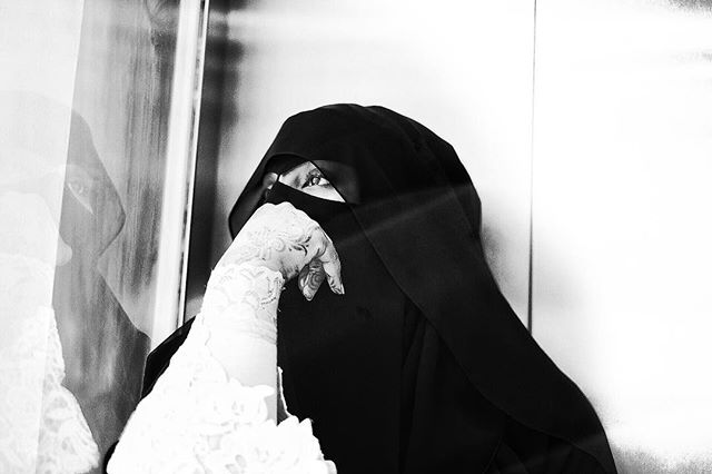 A woman wearing a black niqab with a patterned white glove poses in a contemplative, black and white image.