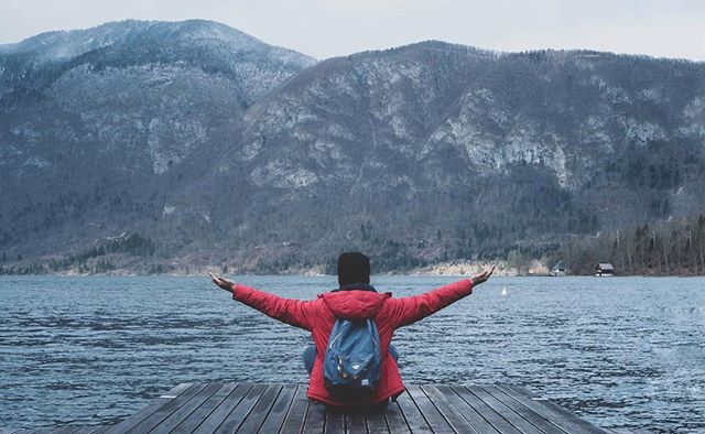 Man with backpack sits at the end of a wooden dock enjoying the mountain lake view.