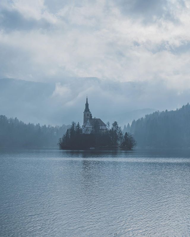 A serene view of Lake Bled with a church atop a small island, shrouded in mist and surrounded by mountains.