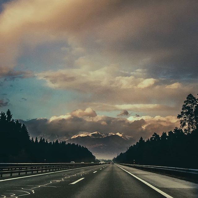 A scenic highway stretches towards distant mountains under a cloudy sky, surrounded by lush trees, evoking a sense of adventure.