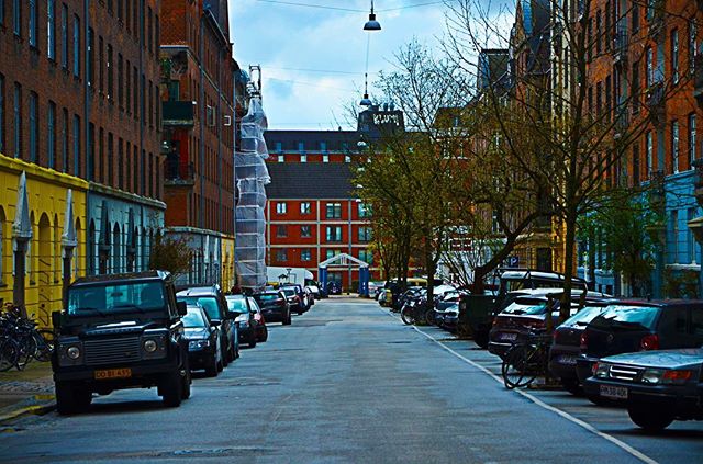 A quiet city street scene with parked cars, brick buildings, and scaffolding-covered structure.