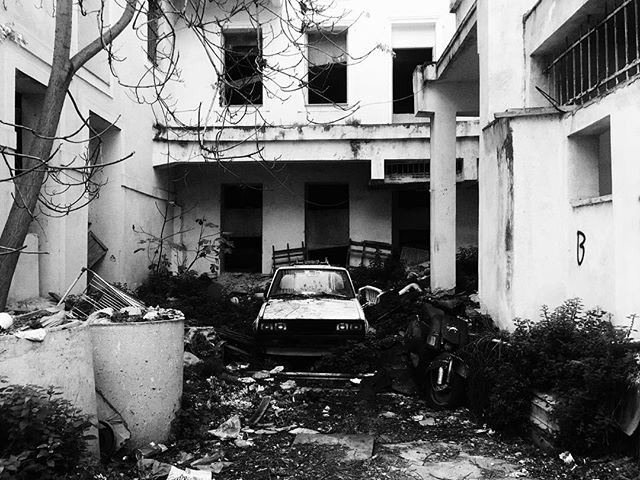 An abandoned car and motorcycle sit in the overgrown courtyard of a decaying building in stark black and white.