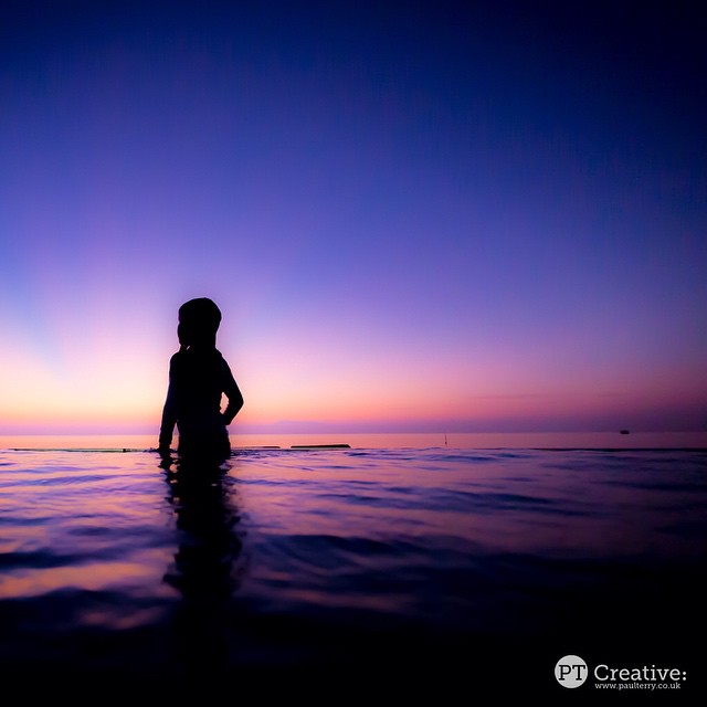 A child's silhouette stands peacefully in an infinity pool during a vibrant sunset by the ocean.
