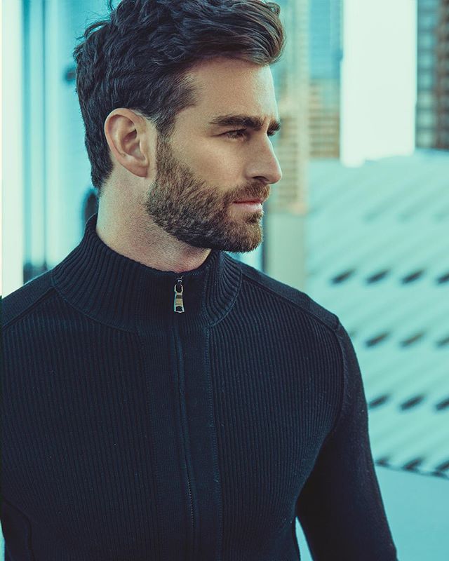 Portrait of a bearded man in a black sweater, looking confident in an urban setting.