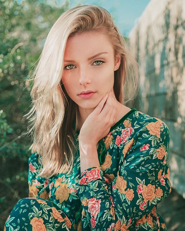 A blonde woman with green eyes wearing a floral dress poses softly in natural light.
