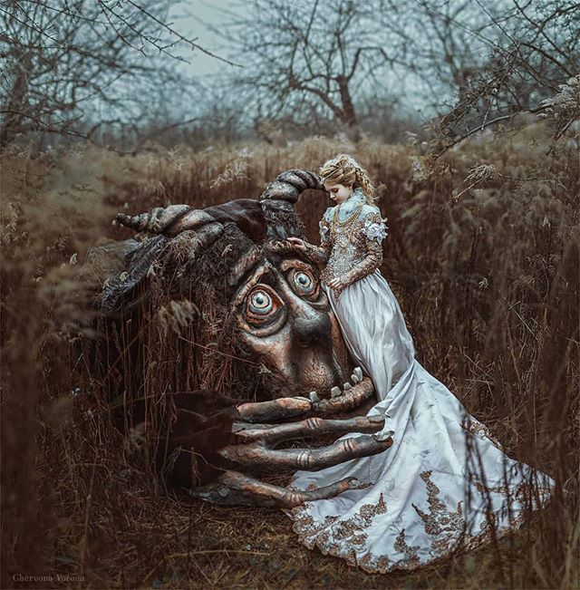 A woman in a gown stands next to a large, sad fantasy creature in a field.