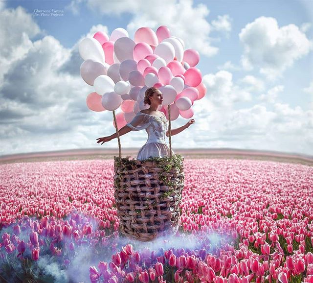A woman floats above a field of pink tulips in a balloon basket under a cloudy sky.