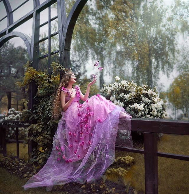 A woman in a pink gown sits amongst foliage, looking up at floating butterflies in a dreamy garden setting.