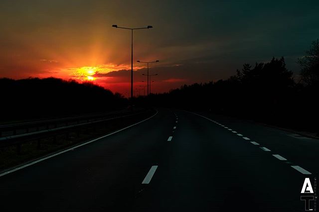 An open road at sunset, lined with street lights, fades into the horizon, creating a dramatic scene.