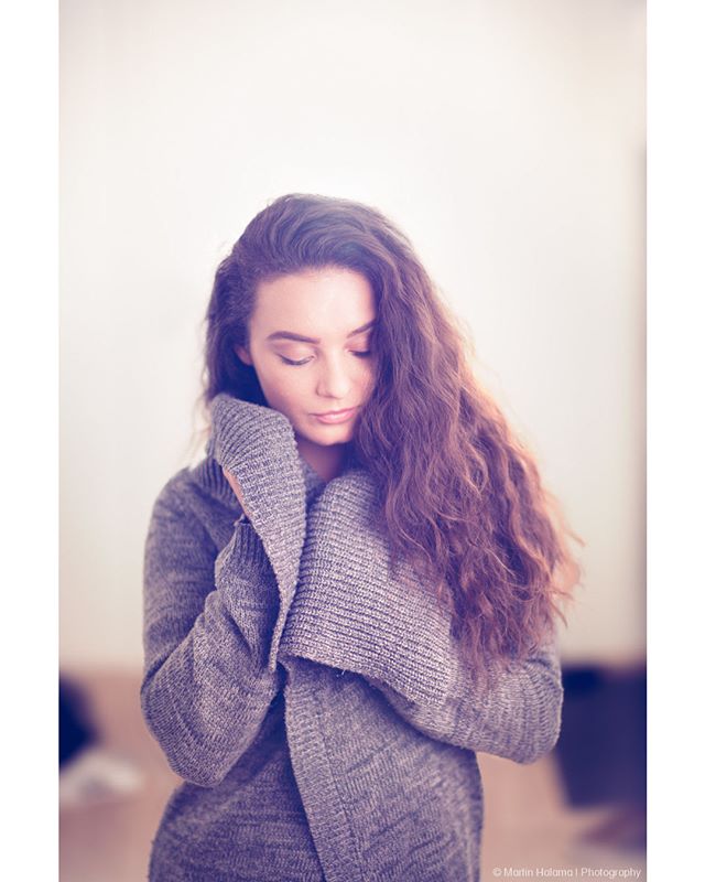 A woman with curly hair looks down in a soft, contemplative portrait indoors.
