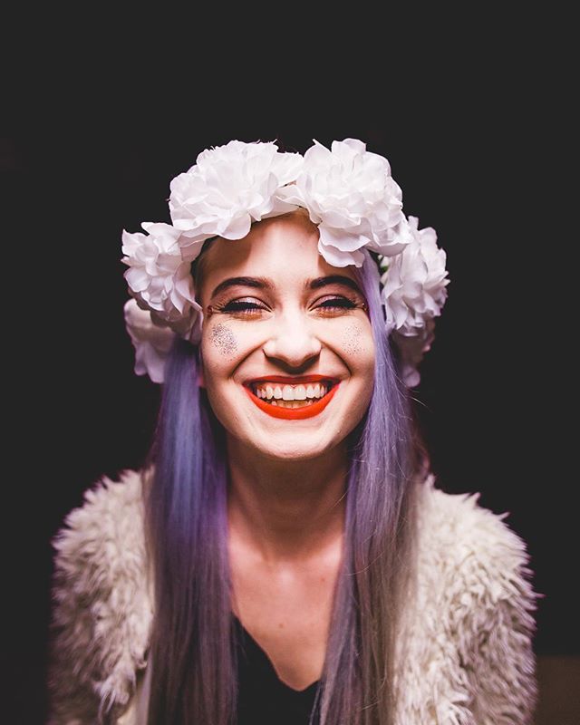A cheerful woman with purple hair and a flower crown smiles against a dark backdrop.