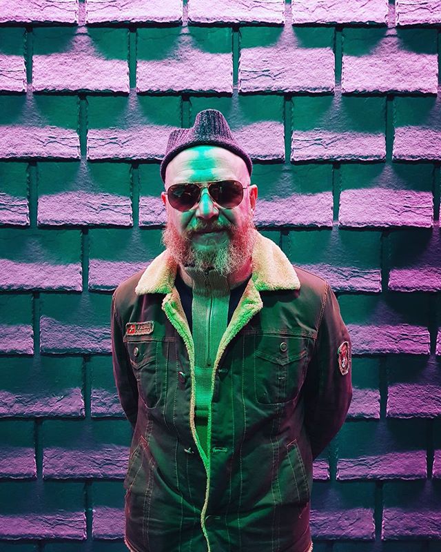 A stylish man with a beard and sunglasses poses in front of a textured wall with unique, moody lighting.