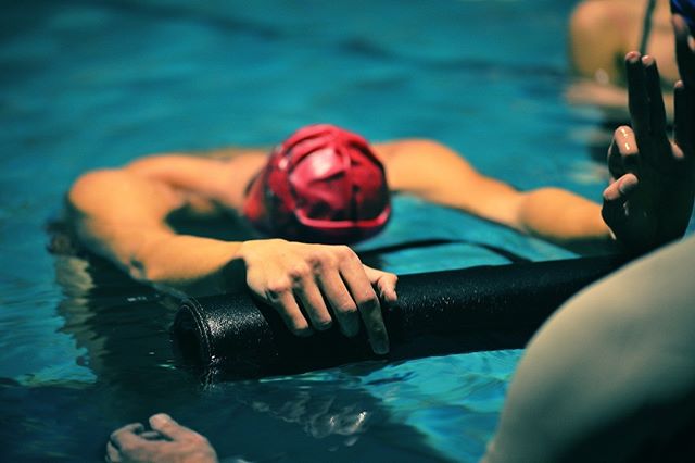 A swimmer rests in a pool, holding onto a black floatation device, training or recovering. 