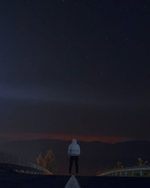 A lone figure stands on a dark road under a starry sky, facing distant mountains at night.