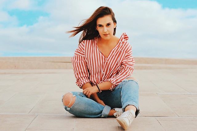 A woman in a red striped shirt and ripped jeans sits cross-legged outdoors.