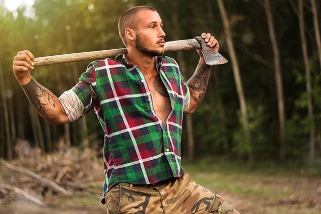 A muscular man with tattoos carries an axe over his shoulder in a forest setting.