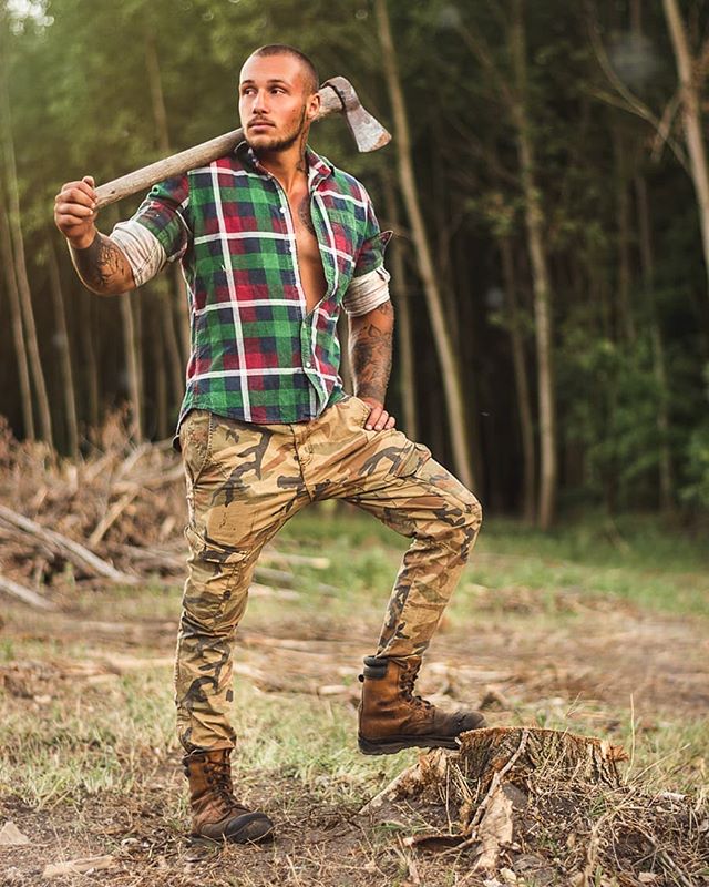 A muscular man in a flannel shirt and camo pants poses with an axe in a forest.