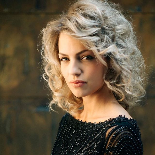 A blonde woman with curly hair poses elegantly in a close-up portrait against a warm backdrop.