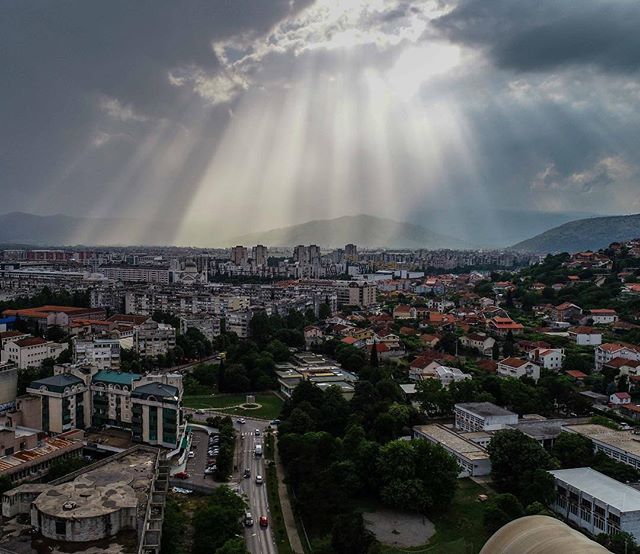 A cityscape view featuring buildings, houses, and mountains under sun rays piercing through the clouds.