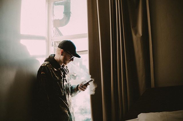 A man in a jacket and cap leans against a wall by a window, vaping and looking at his phone.