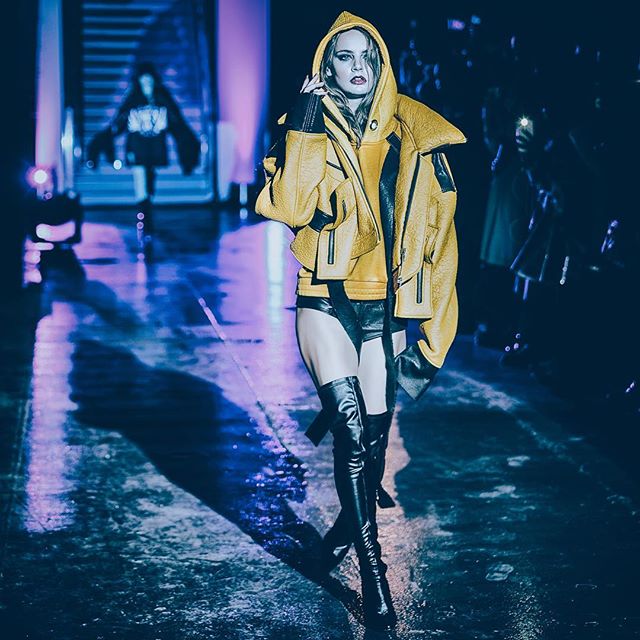 A confident fashion model strides down the runway wearing a yellow hooded jacket and thigh-high boots.