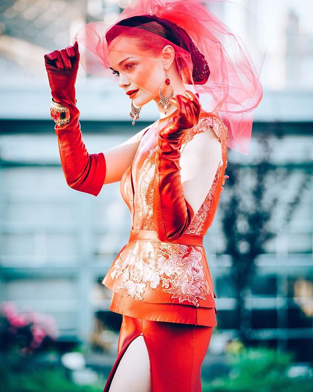A woman in a red dress, hat, and gloves poses for a fashion editorial.