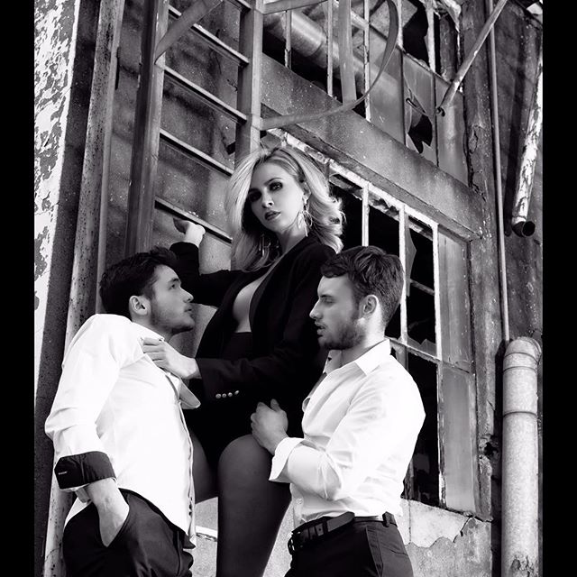 A dramatic black and white image of a woman and two men posing together in front of an industrial building.