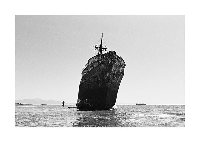 Monochrome shot of a man walking towards a large shipwreck in the sea under a clear sky.