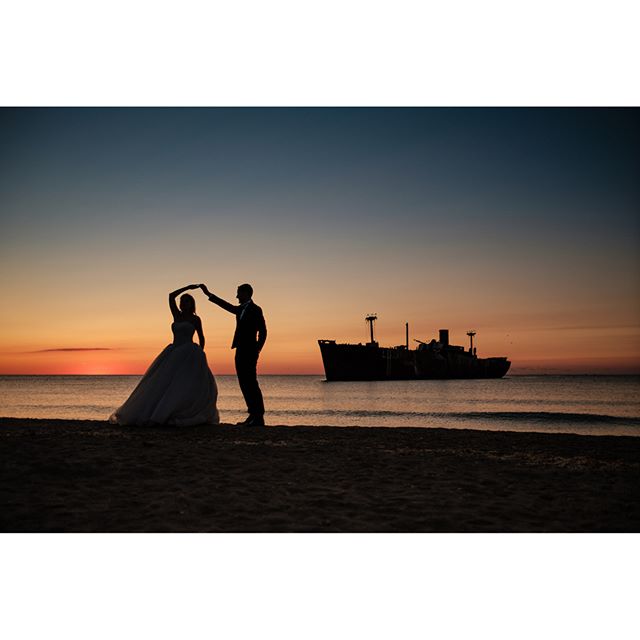 A bride and groom silhouetted against a sunset with a shipwreck in the background.