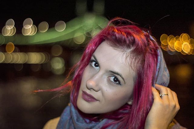 A woman with pink hair poses against a blurred background of city lights at night.