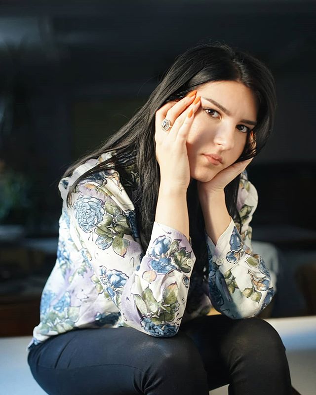 A young woman with dark hair rests her face in her hands, looking thoughtfully at the camera.