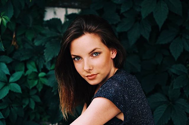 A serene woman with brunette hair poses naturally against a leafy green backdrop in soft daylight.