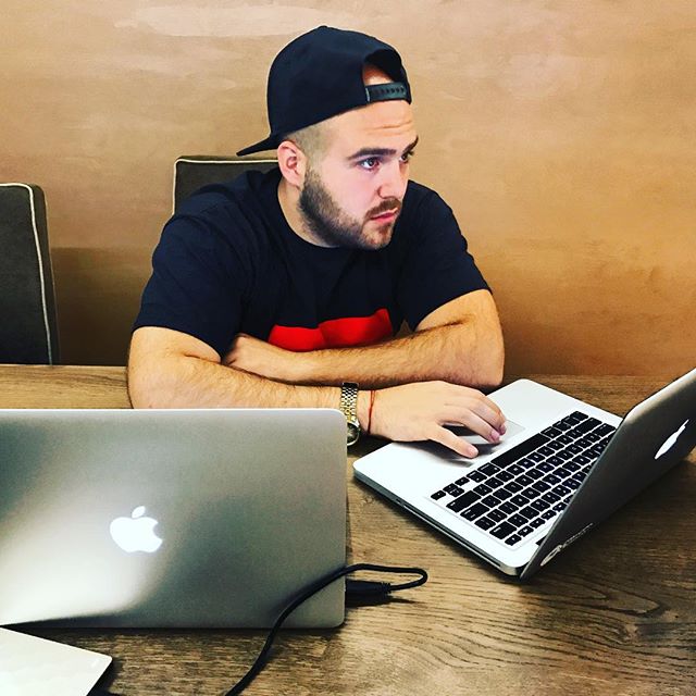 A man working on two laptops at a wooden table, focused on his task.