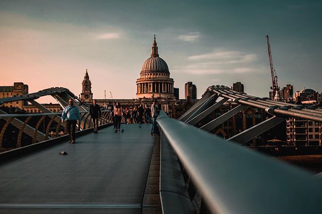 People walk across Millennium Bridge in London with St. Paul's Cathedral in the background during a tranquil sunset.