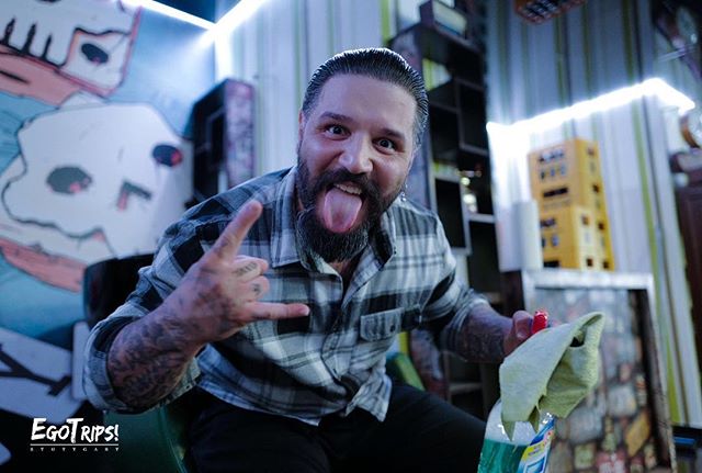 A bearded man playfully sticks out his tongue and makes a rock and roll sign in a studio setting with wall art.