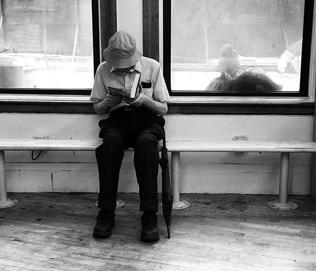 A man in a hat sits on a bench, engrossed in a book, with an umbrella beside him.