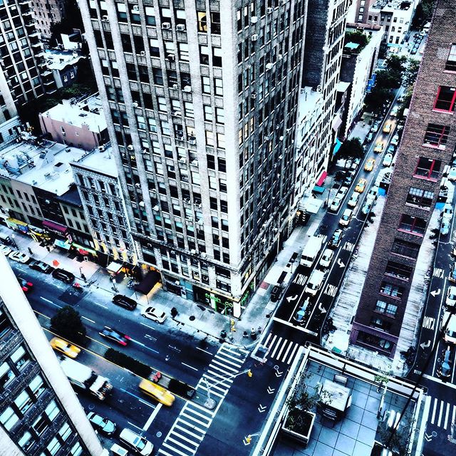 An aerial view captures a busy New York street filled with cars and taxis between tall buildings.
