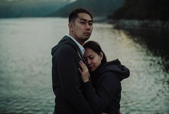 A couple embrace outdoors by a lake with mountains in the background.