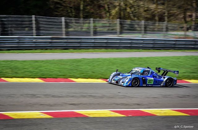 A blue race car speeds along a racetrack on a sunny day, showcasing the thrill of motorsport.