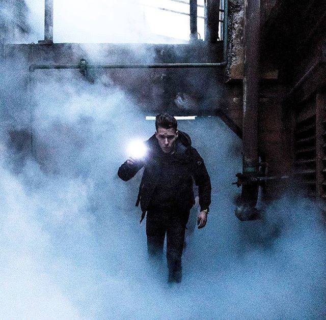 A man walks through a smoky, industrial alleyway holding a flashlight, creating a mysterious and suspenseful atmosphere.