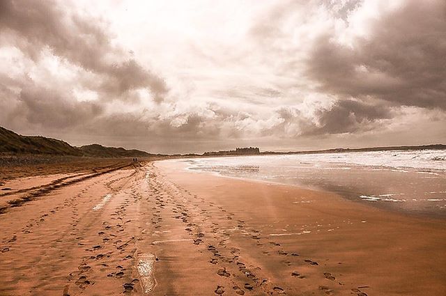 A serene beach with footprints in the sand, under a cloudy sky. Calm ocean waves meet the shore.