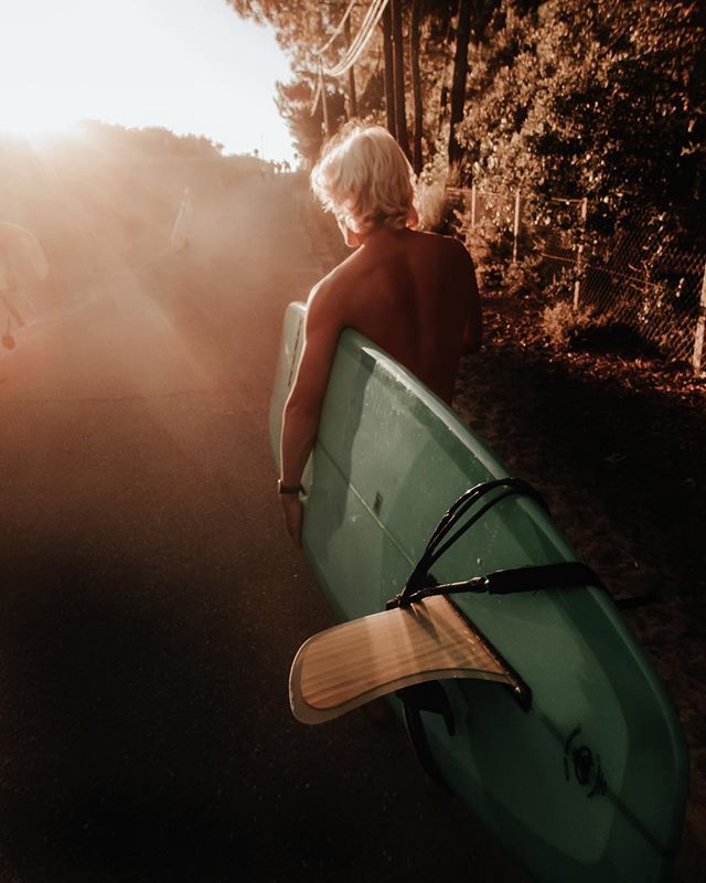 A man with a surfboard walks down the road in the sunlight.