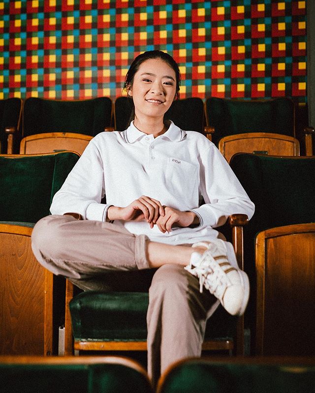 A smiling young woman sits in a theater chair wearing casual clothes and sneakers.