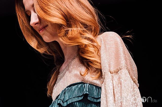 A redhead woman in an elegant outfit poses in soft light against a black background.