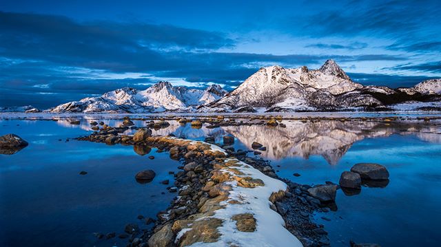 Snowy mountains reflecting in still water create a peaceful winter landscape at dusk.