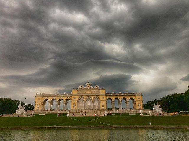 Schönbrunn Palace in Vienna, Austria stands majestic under a dramatic cloudy sky, reflected in a serene pond.