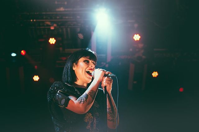 A tattooed female singer performs with energy on a concert stage, illuminated by stage lights.
