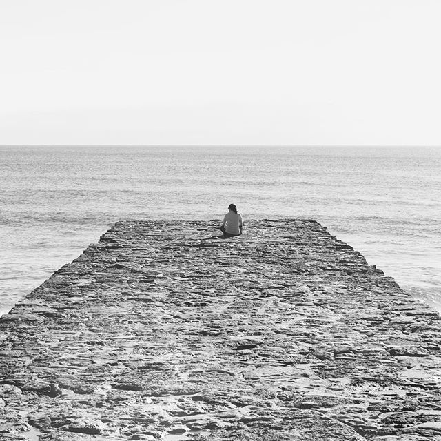 A lone person sits on a stone pier, contemplating the serene ocean view in a calming monochrome scene.